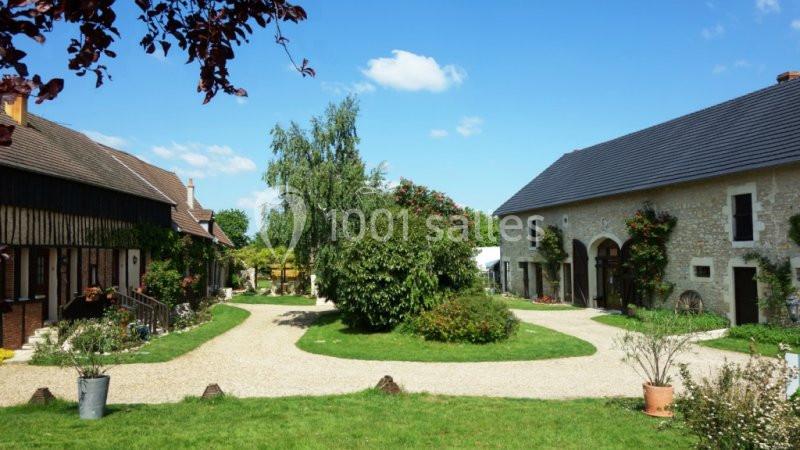 Cour intérieure avec bâtiments en pierre et en bois, entourée de végétation et d'allées gravillonnées sous un ciel bleu.