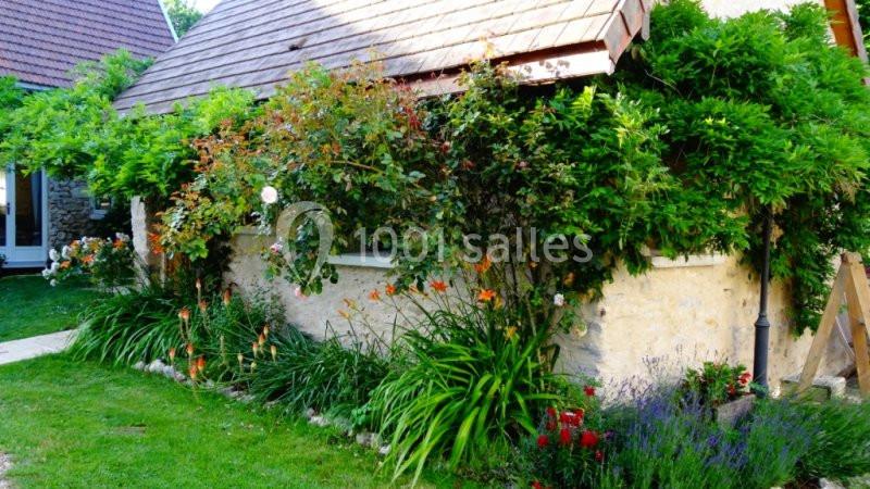 Maison en pierre avec jardin fleuri, entourée de plantes grimpantes et de fleurs colorées, sous un toit en tuiles.