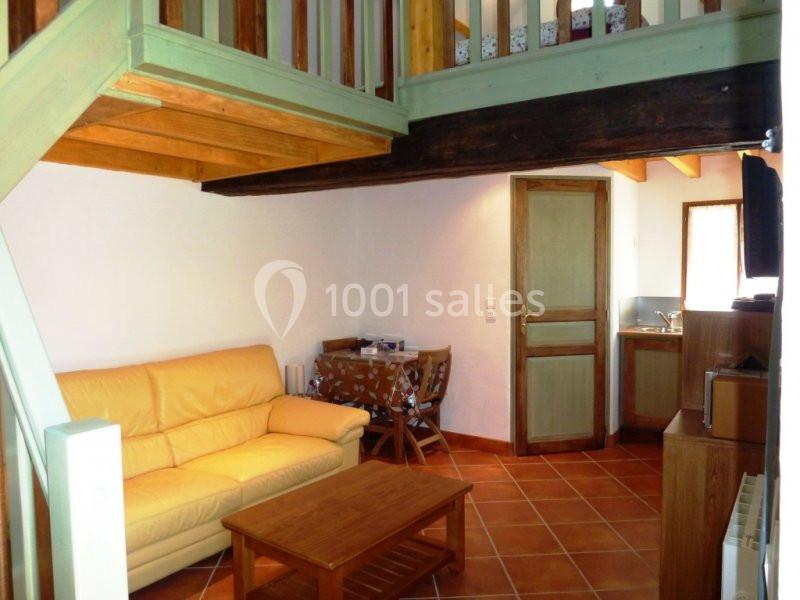 Salon avec canapé jaune, table basse en bois, mezzanine en bois, coin repas et kitchenette dans un intérieur rustique.