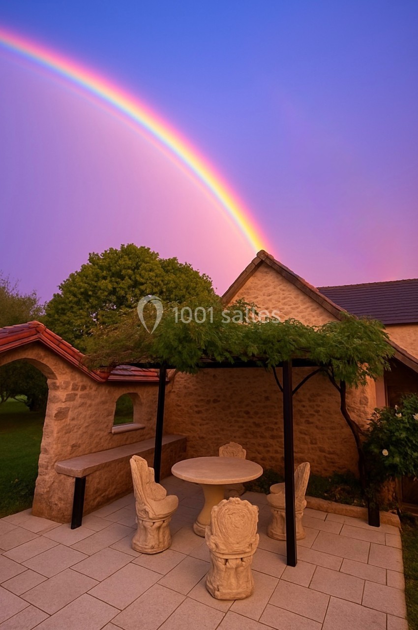 Location salle Beaulieu-sur-Loire (Loiret) - Domaine des Medards #6 Terrasse en pierre avec table et chaises sculptées, pergola végétalisée, arc-en-ciel et ciel violet en arrière-plan.
