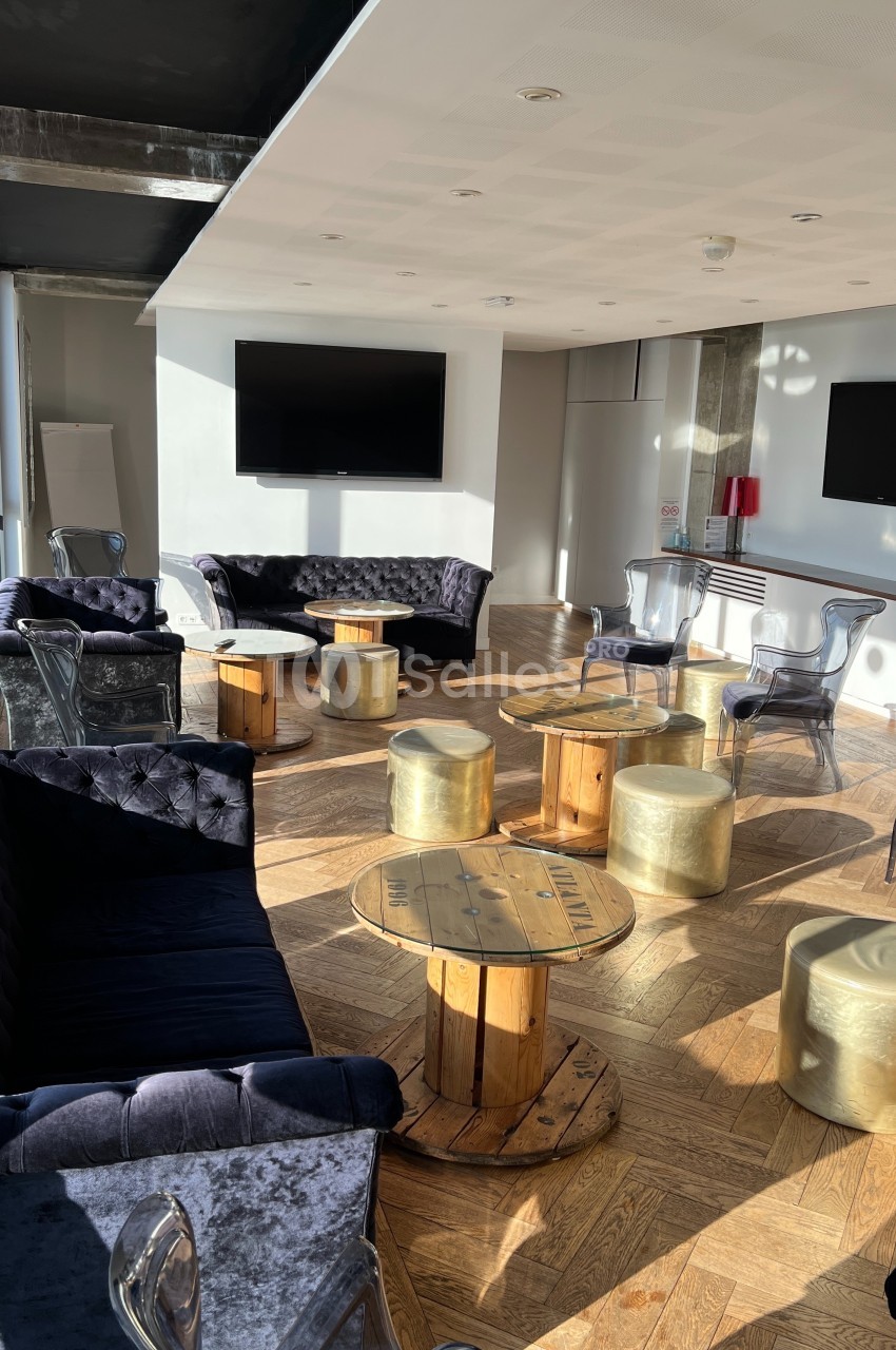 Salle lumineuse avec parquet, canapés en velours, tables en bois et tabourets dorés, éclairée par la lumière naturelle.