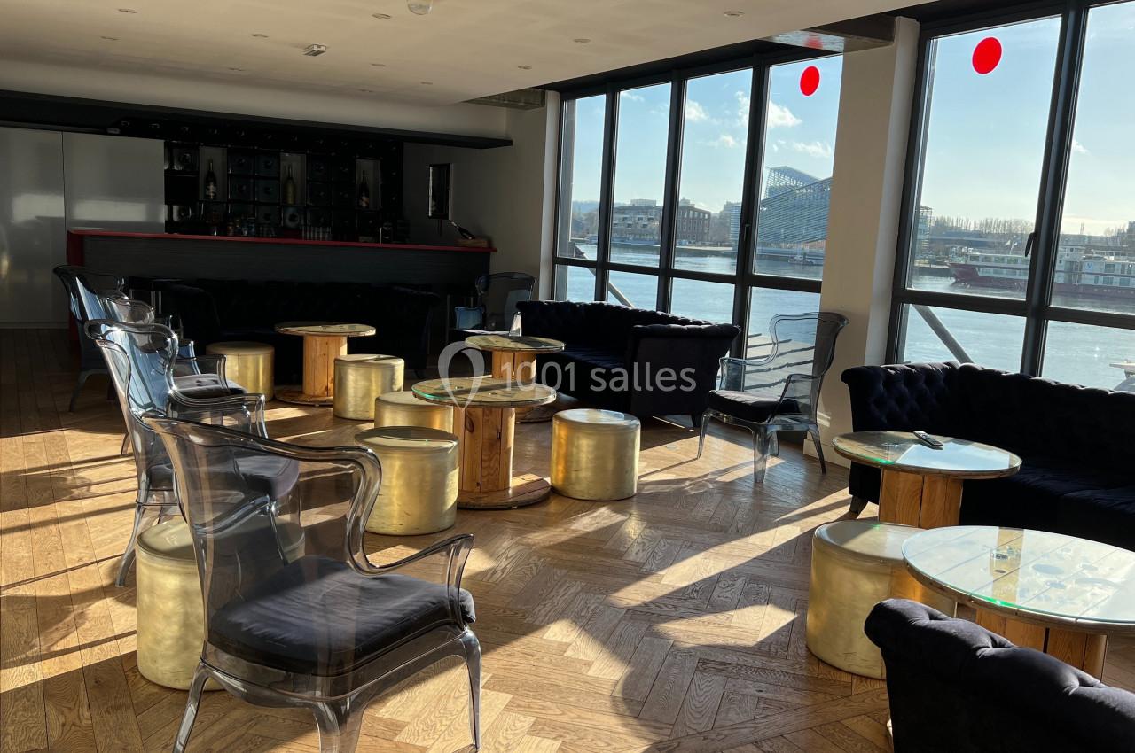 Salle lumineuse avec grandes baies vitrées, mobilier élégant en bois et métal, vue sur un fleuve et des bâtiments.