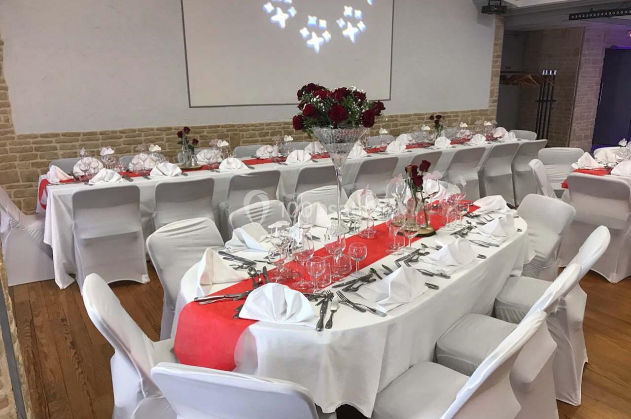 Salle décorée pour un repas avec tables dressées, nappes blanches, chemins de table rouges et compositions florales.