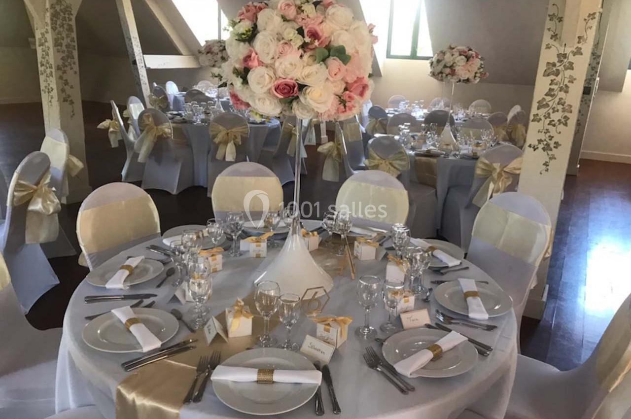 Salle de réception décorée avec des tables rondes, nappes blanches, chaises ornées de nœuds dorés et centre de table floral.