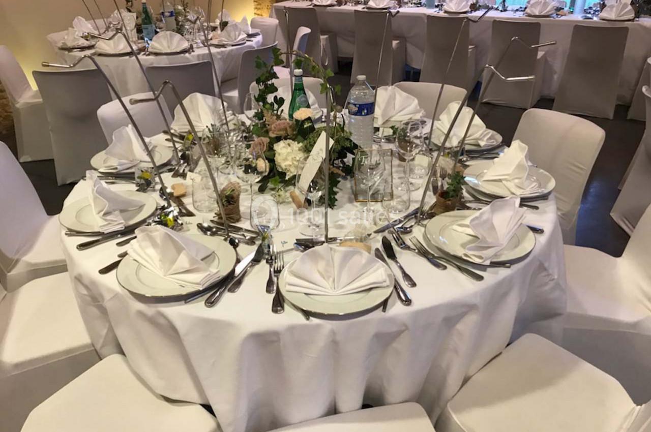 Table ronde élégamment dressée avec nappes blanches, serviettes pliées, couverts disposés et décoration florale au centre.