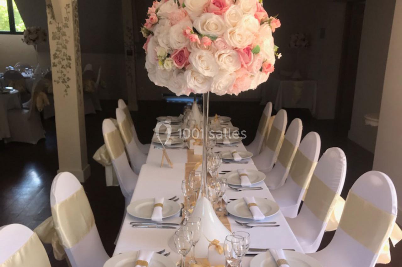 Table décorée pour un événement, avec nappes blanches, chaises ornées de rubans dorés et centre de table floral.