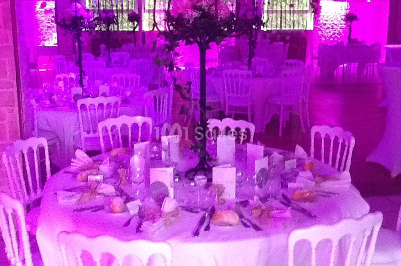 Salle de réception décorée avec des tables rondes dressées, éclairage rose et chaises blanches.
