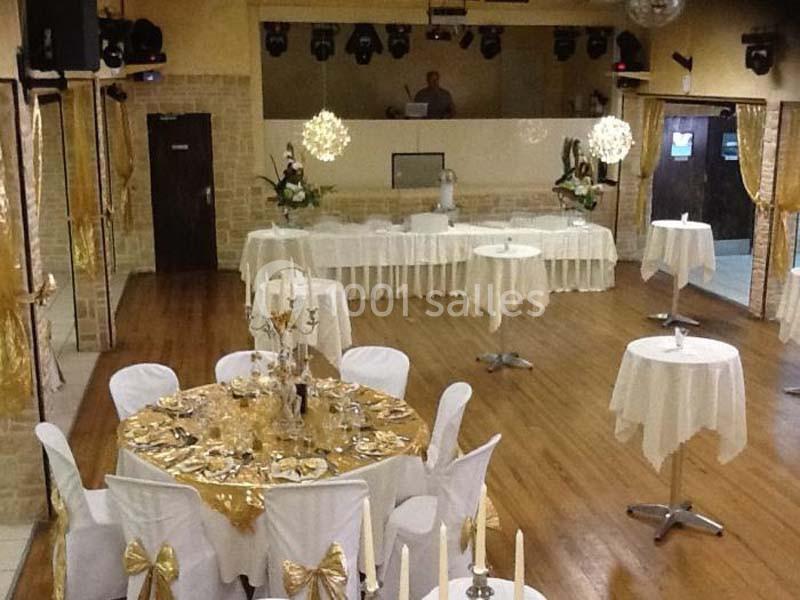 Salle de réception décorée avec des nappes blanches et dorées, tables rondes et hautes, et une piste de danse au fond.