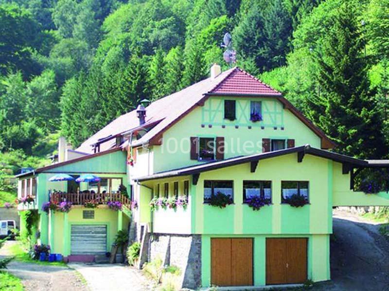 Maison à deux étages avec façade verte, balcons fleuris et entourée de forêt dense.