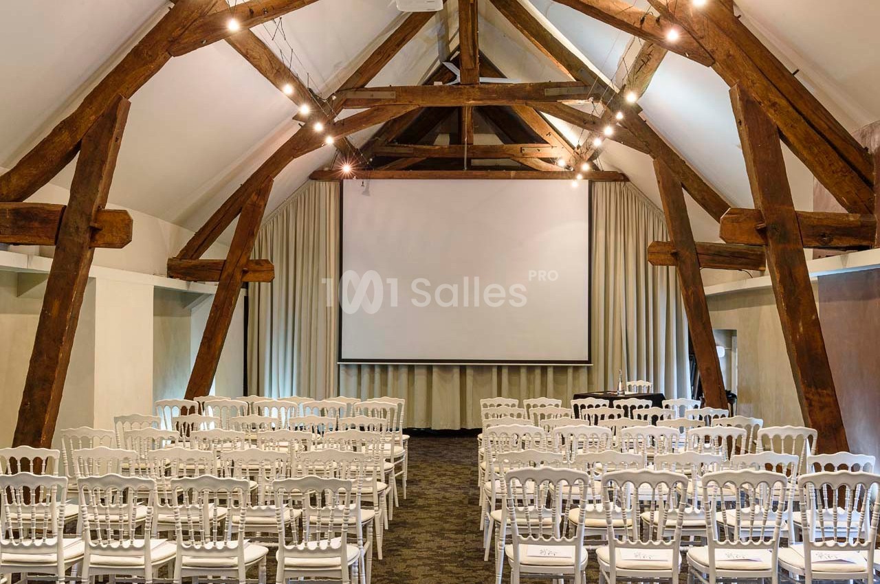 Salle de conférence avec poutres apparentes, écran de projection, rideaux beiges et chaises blanches alignées.
