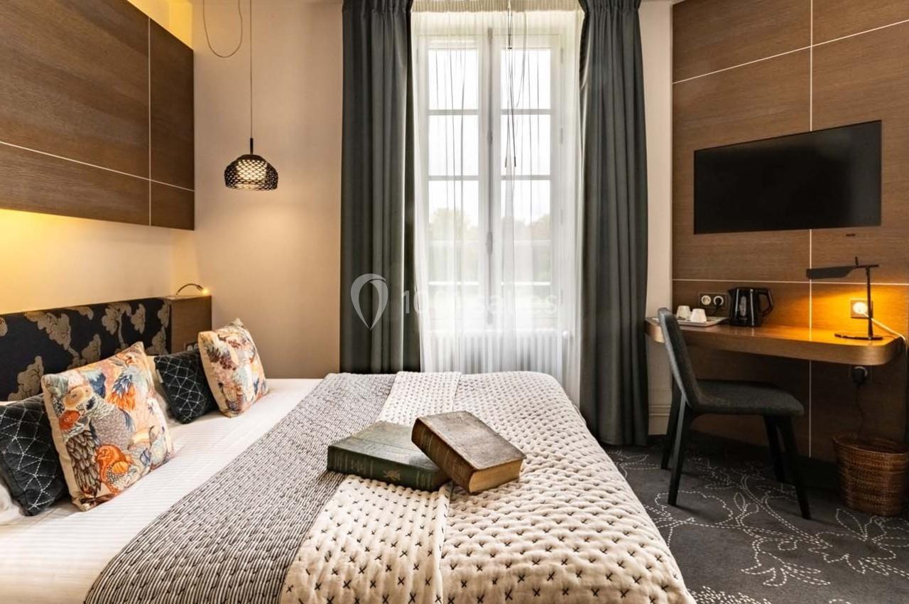 Chambre d'hôtel lumineuse avec lit double, coussins décoratifs, bureau, télévision murale et fenêtre avec rideaux.