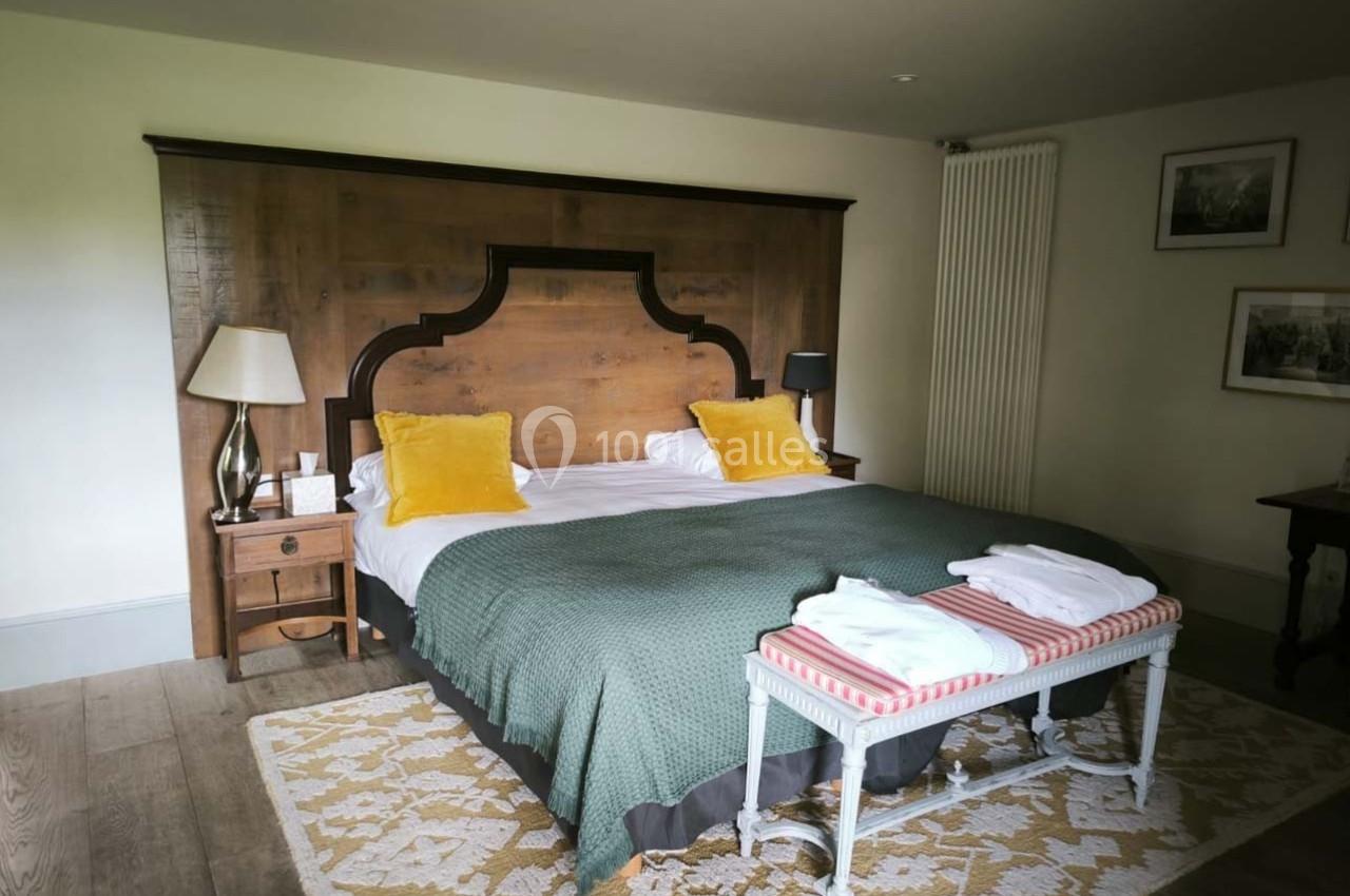 Lit double avec tête de lit en bois, coussins jaunes, plaid vert, banc avec serviettes pliées, dans une chambre lumineuse.