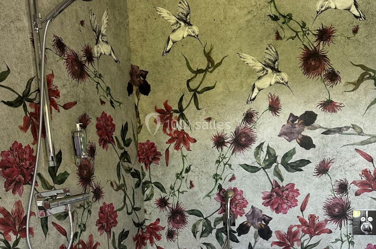 Mur de salle de bain décoré de motifs floraux rouges et verts avec des oiseaux blancs, douche et baignoire visibles.