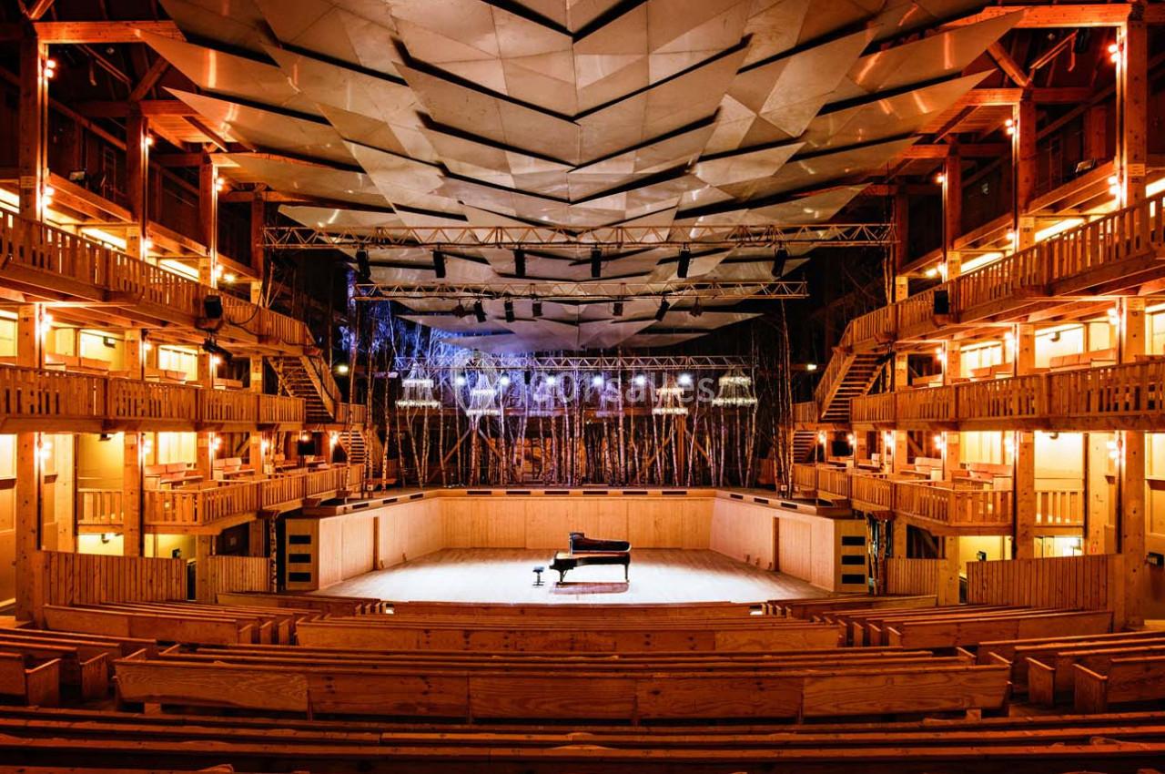 Salle de concert en bois avec un piano à queue sur scène, éclairée par des lumières chaudes et un plafond géométrique.