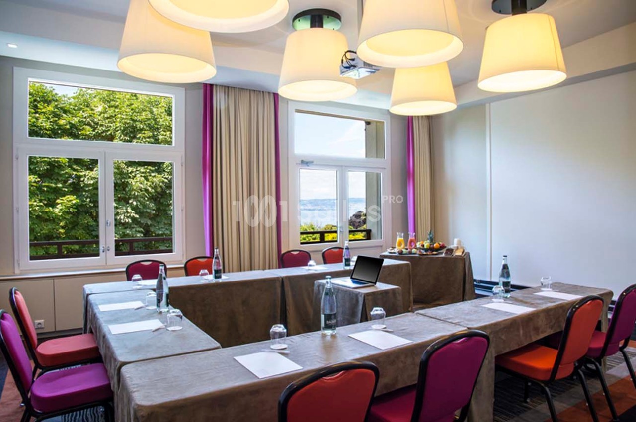 Salle de réunion lumineuse avec tables en U, chaises rouges, grandes fenêtres donnant sur un paysage verdoyant et un lac.