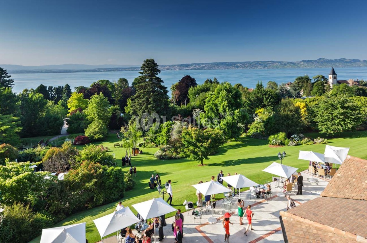 Jardin verdoyant avec vue sur un lac, des montagnes à l'horizon et des parasols blancs sur une terrasse animée.