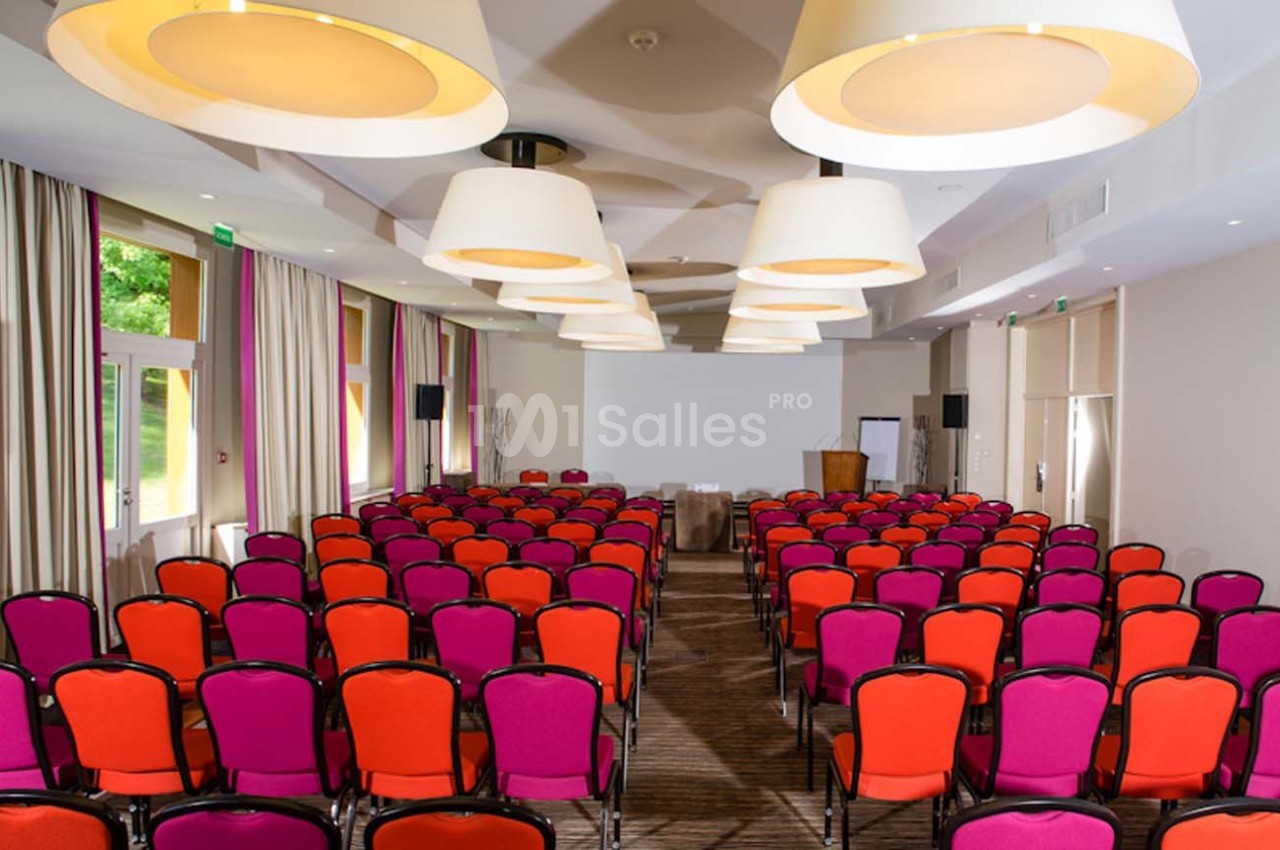 Salle de conférence avec des rangées de chaises rouges et violettes, éclairée par des suspensions lumineuses.
