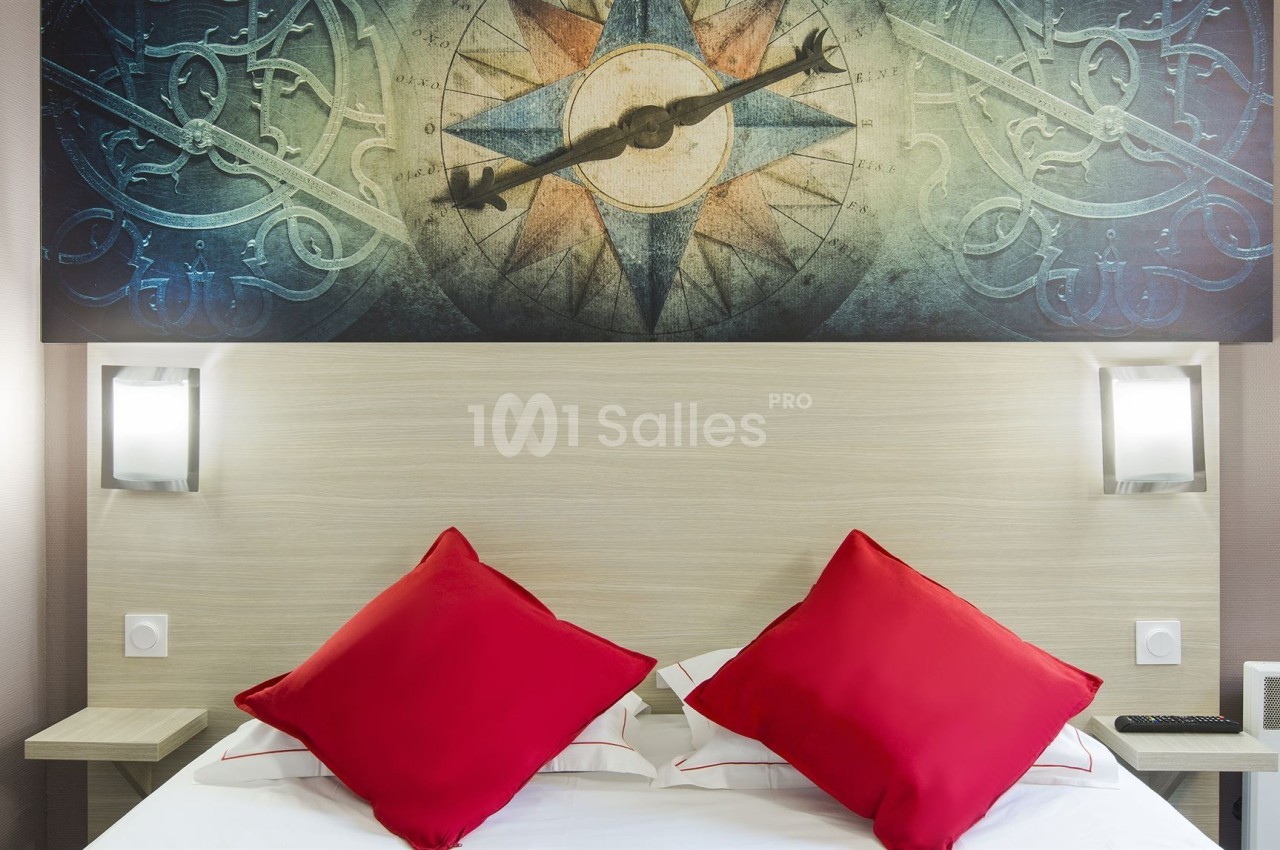 Lit avec deux coussins rouges, encadré par deux lampes murales, sous un tableau représentant une boussole.