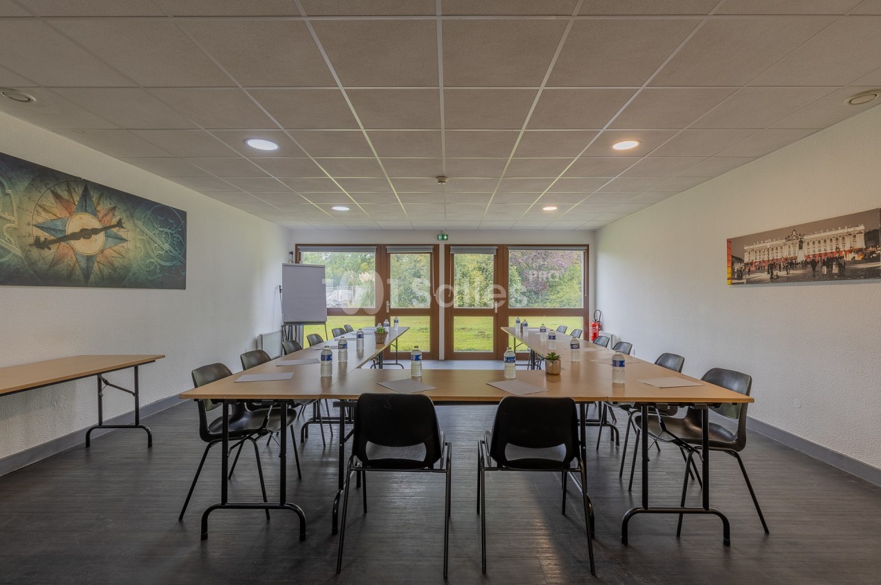 Salle de réunion lumineuse avec tables en U, chaises noires, bouteilles d'eau et vue sur un espace vert à travers des baies…