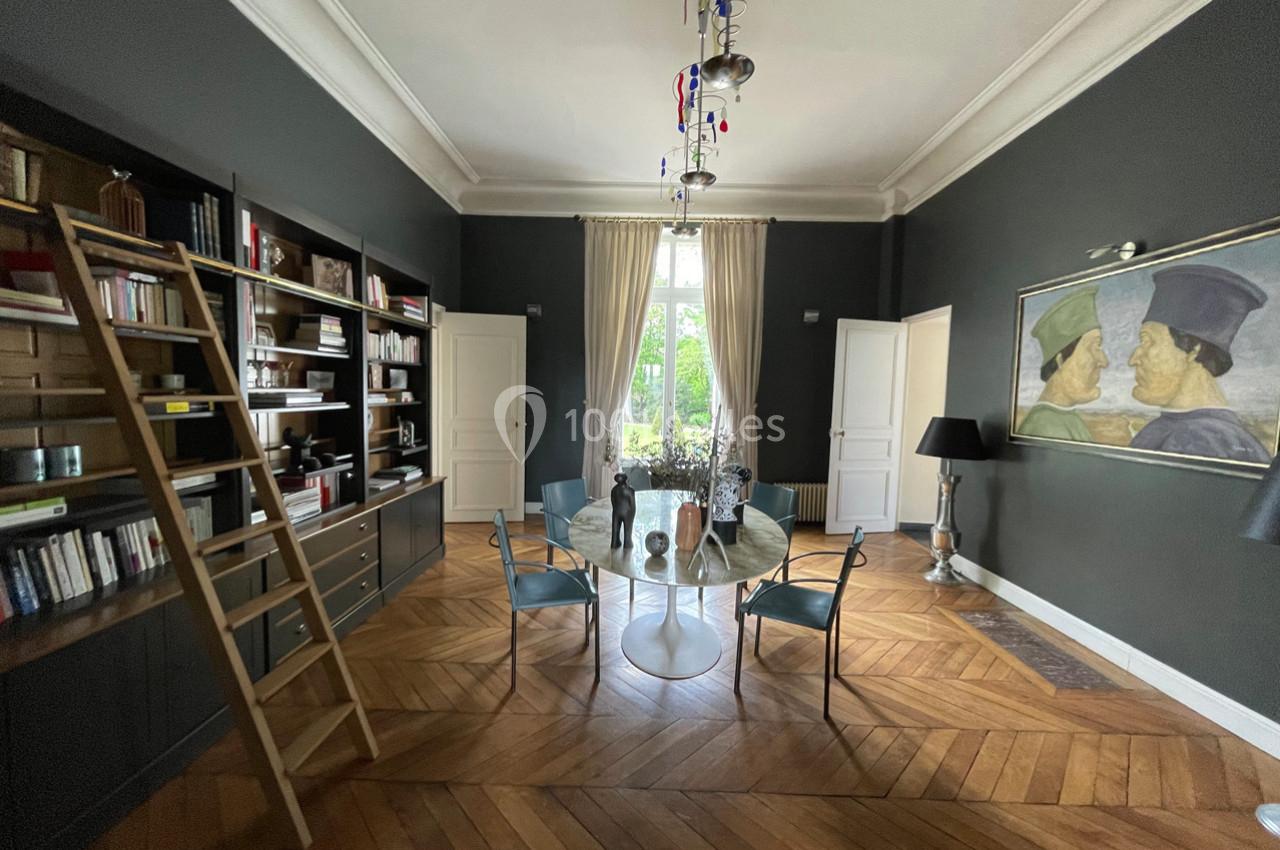 Salle à manger avec parquet en bois, table ronde, chaises, bibliothèque avec échelle et grande fenêtre donnant sur un jardin.