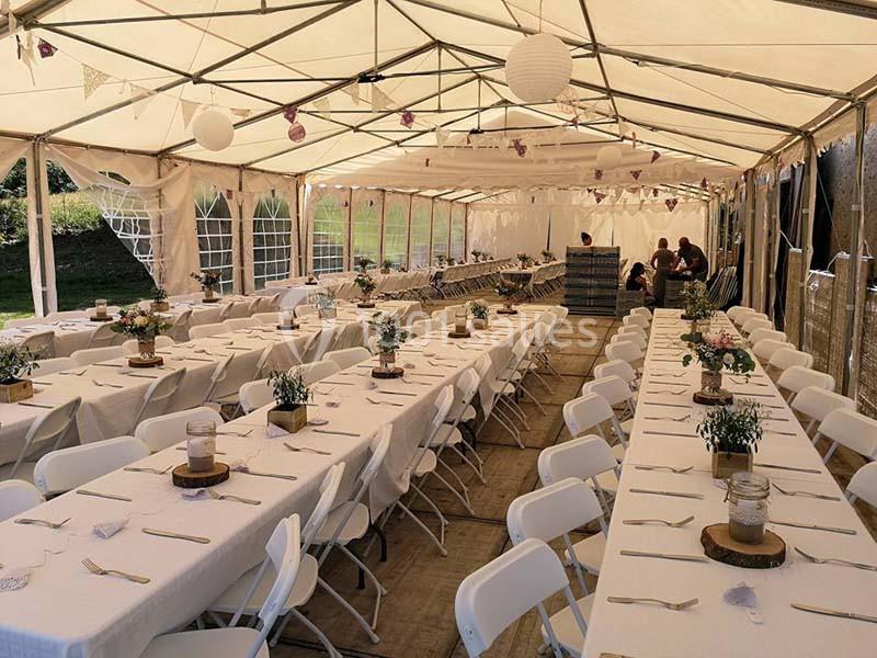Intérieur d'une tente de réception avec de longues tables dressées, décorées de fleurs et de lanternes suspendues.