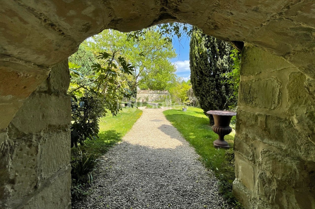 Vue d'un jardin verdoyant à travers une arche en pierre, avec un chemin de gravier et des arbres en arrière-plan.