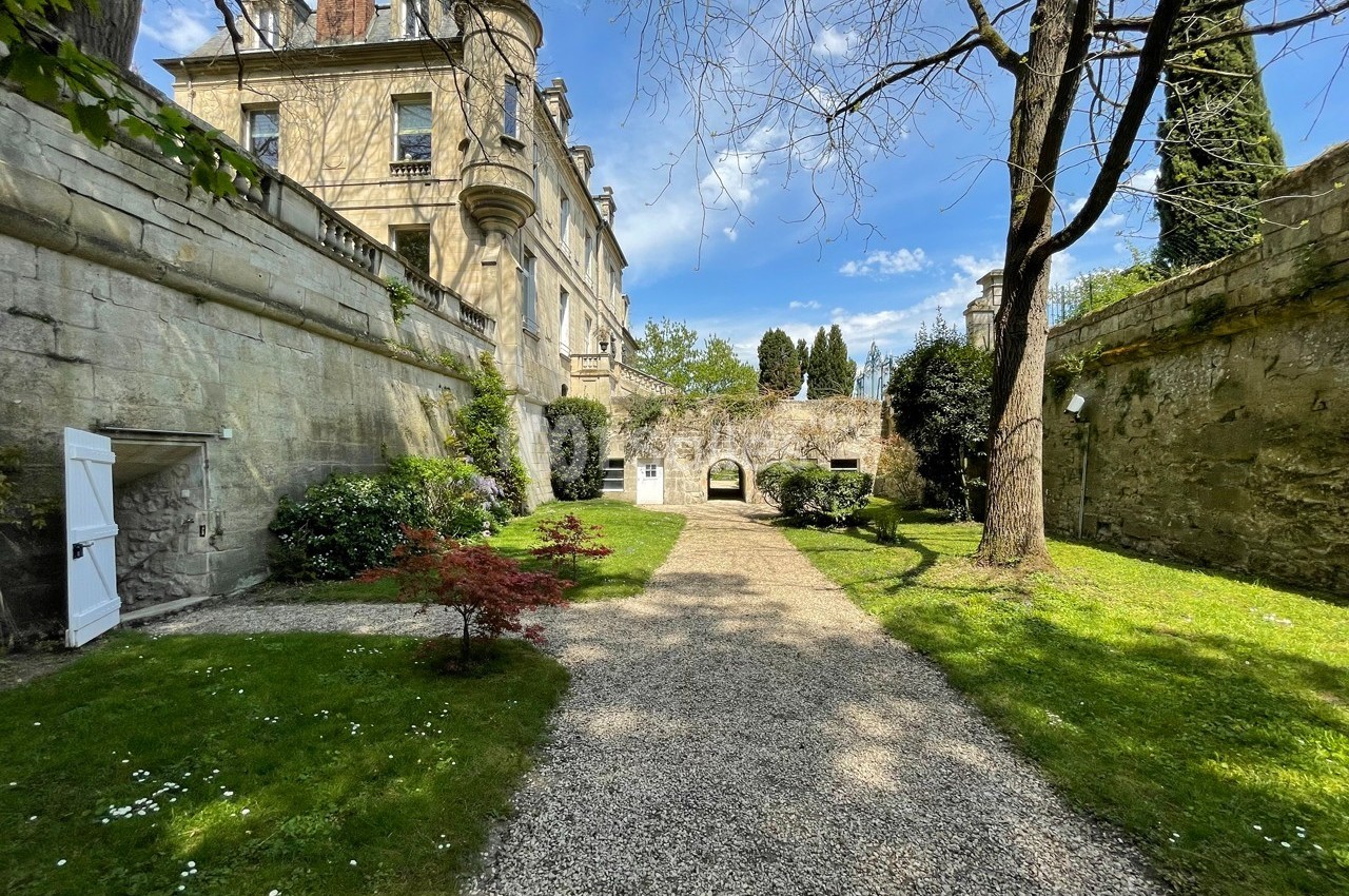 Allée gravillonnée bordée de pelouses, arbres et arbustes, menant à une bâtisse en pierre sous un ciel dégagé.