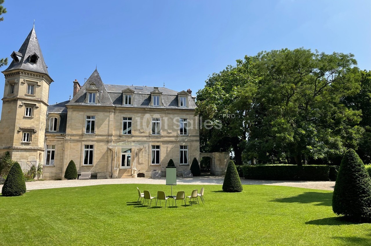 Façade d’un château en pierre entouré d’un jardin avec pelouse, chaises disposées en cercle et arbres en arrière-plan.