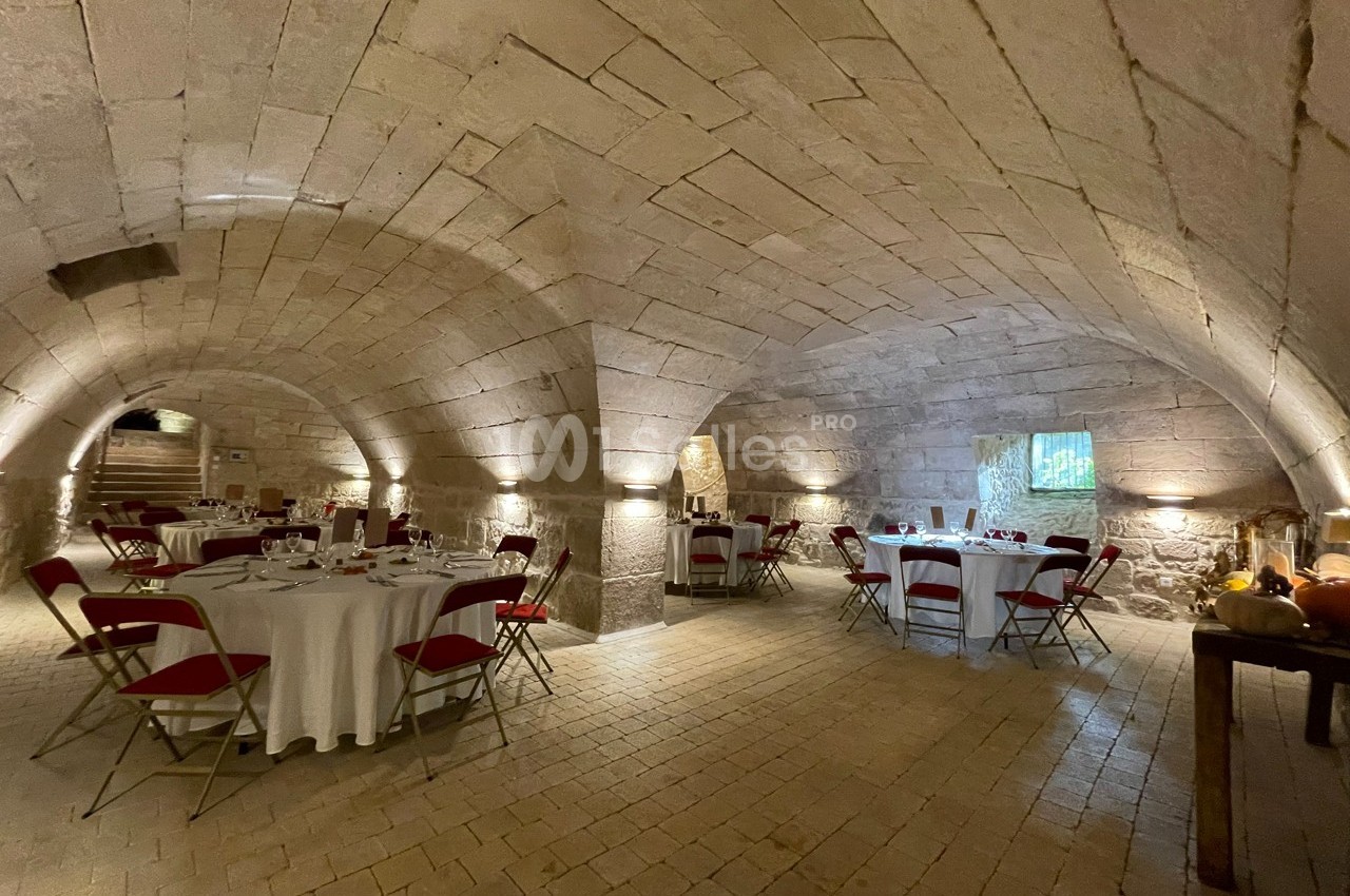Salle voûtée en pierre avec tables rondes dressées, chaises rouges et éclairage tamisé.