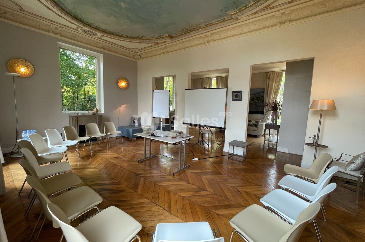 Salle lumineuse avec parquet, chaises disposées en cercle, tableaux blancs et projecteur, entourée de grandes fenêtres.