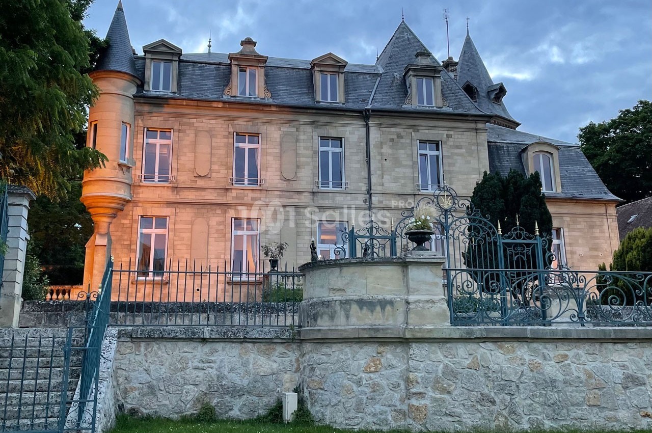 Façade d'un bâtiment en pierre de style classique avec tourelle, entouré d'une grille en fer forgé et d'un jardin.