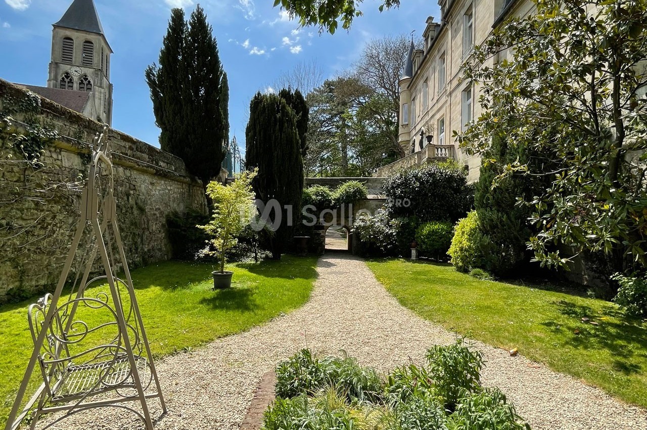 Jardin avec allée gravillonnée, bancs, arbres et façade de bâtiment ancien sous un ciel ensoleillé.