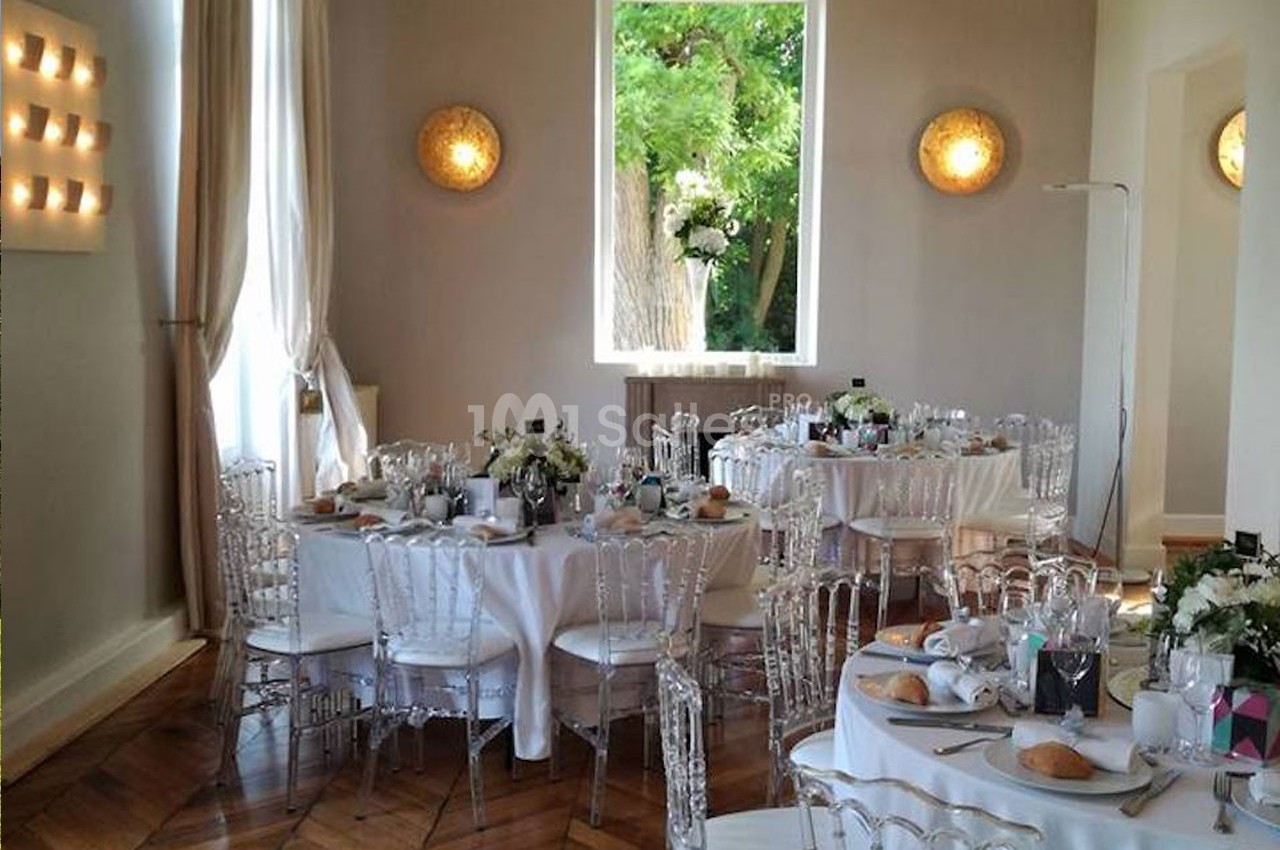 Salle de réception lumineuse avec tables rondes dressées, chaises transparentes et grande fenêtre donnant sur un jardin…