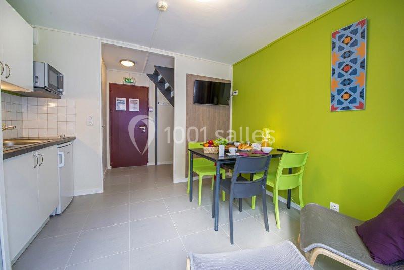 Cuisine ouverte avec table à manger colorée, chaises vertes et noires, mur vert vif et porte d'entrée en arrière-plan.