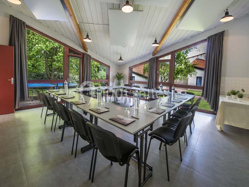 Salle de réunion lumineuse avec de grandes fenêtres donnant sur un jardin, tables disposées en U avec fournitures.