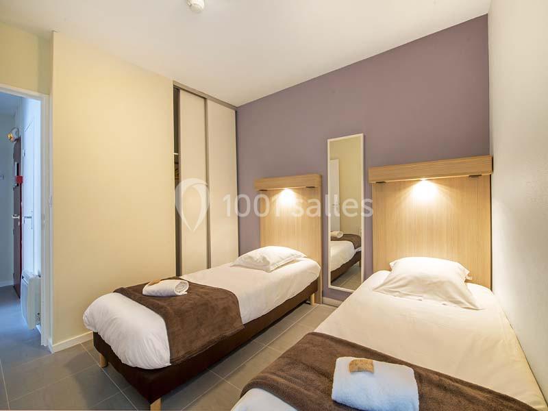 Deux lits simples avec linge de lit brun et blanc, éclairés par des appliques murales dans une chambre lumineuse.