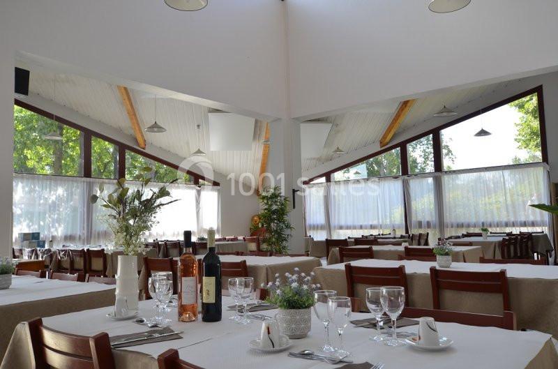 Salle de restaurant lumineuse avec tables dressées, nappes blanches, verres, bouteilles de vin et vue sur l'extérieur…