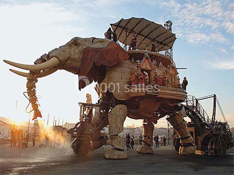 Éléphant mécanique géant en bois et métal transportant des passagers, entouré de visiteurs sur une place.