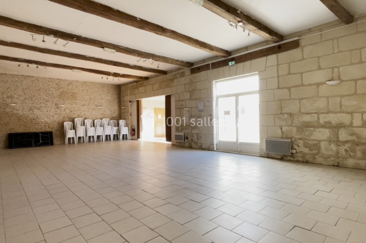 Salle vide avec murs en pierre, poutres apparentes, sol carrelé, chaises empilées et lumière naturelle entrant par une porte…