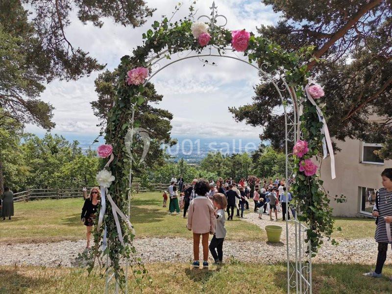Arche fleurie en métal blanc décorée de roses, avec des personnes rassemblées dans un jardin en arrière-plan.