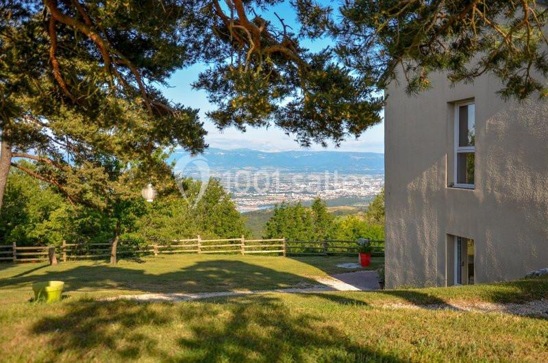 Vue sur un jardin verdoyant avec une maison à droite, des arbres au premier plan et une vallée en arrière-plan.