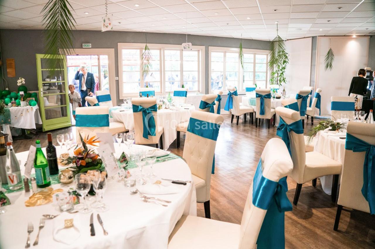 Salle de réception décorée avec des tables rondes, nappes blanches, chaises ornées de nœuds bleus et lumière naturelle.