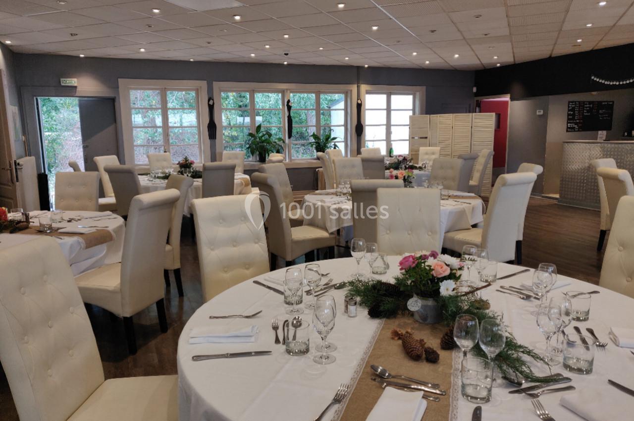 Salle de réception avec tables rondes dressées, nappes blanches, décorations florales et grandes baies vitrées.