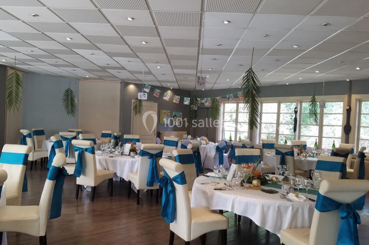 Salle de réception décorée avec des tables rondes, nappes blanches, chaises ornées de rubans bleus et guirlandes suspendues.
