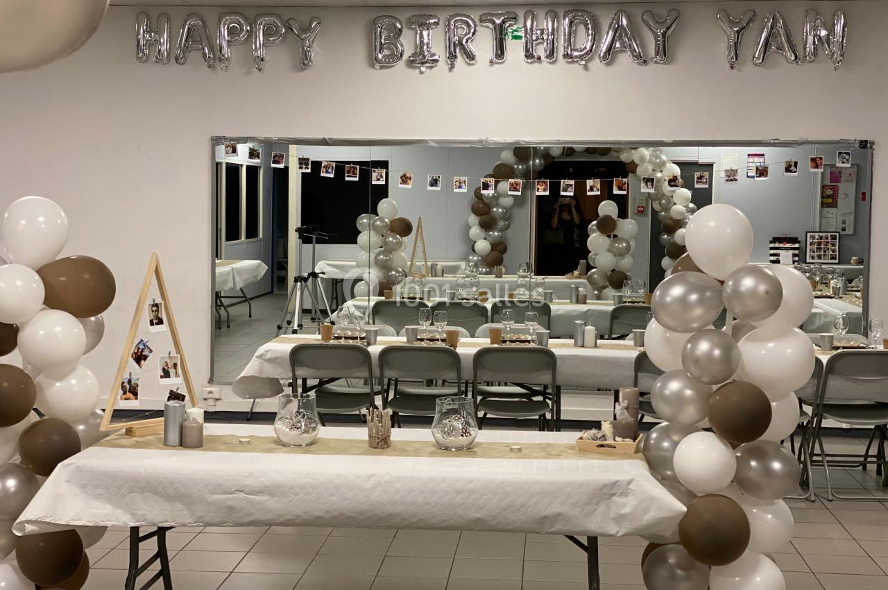 Salle décorée pour un anniversaire avec ballons blancs, marron et argentés, table dressée et guirlande ’Happy Birthday Yan’.