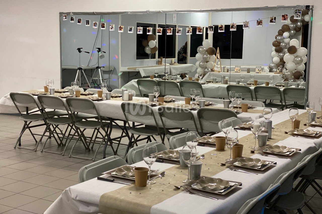 Salle décorée pour une réception avec tables dressées, guirlande de photos et ballons beige et blanc près d'un miroir.