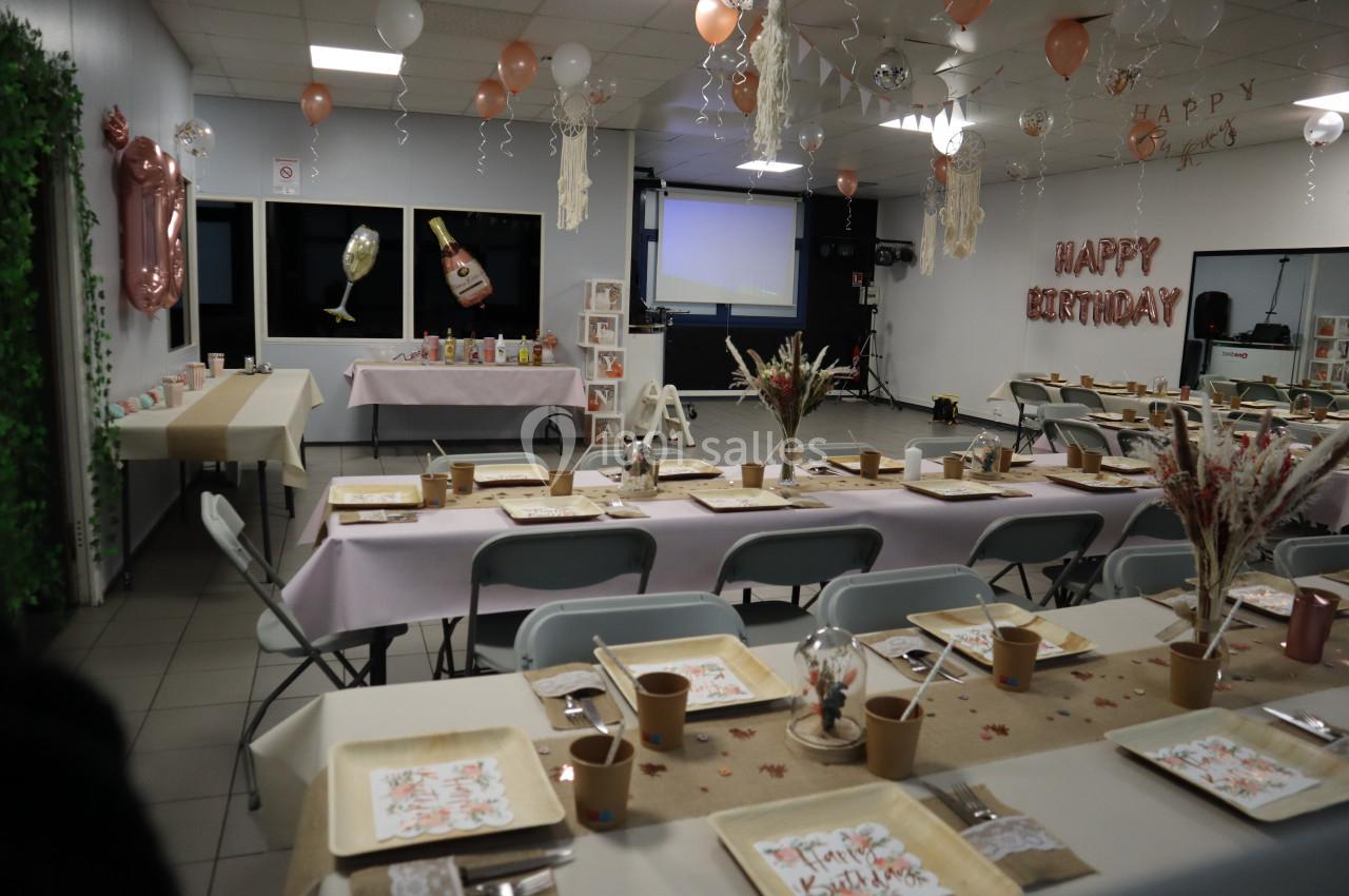 Salle décorée pour un anniversaire avec tables dressées, ballons suspendus et guirlande ’Happy Birthday’ au mur.