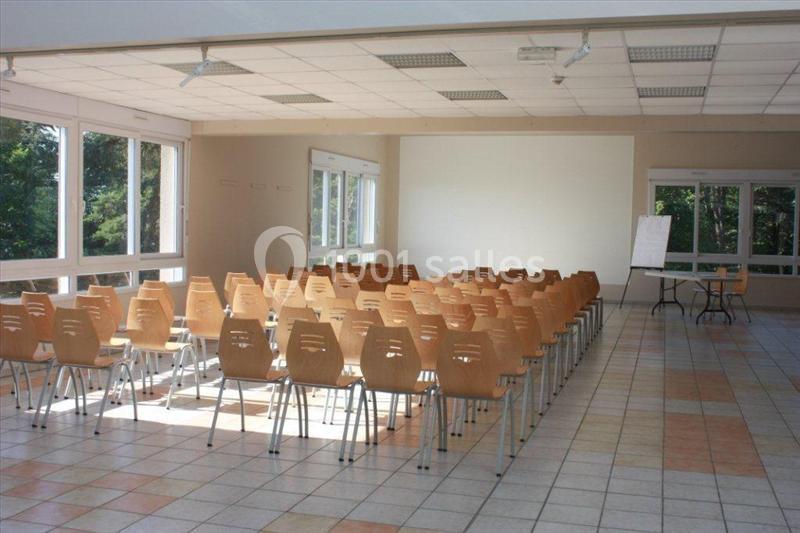 Salle lumineuse avec des rangées de chaises en bois, un tableau blanc et de grandes fenêtres donnant sur un espace vert.