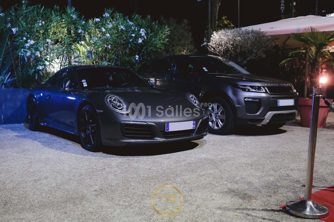 Deux voitures de luxe, une Porsche grise et un SUV noir, stationnées côte à côte dans un espace extérieur éclairé.