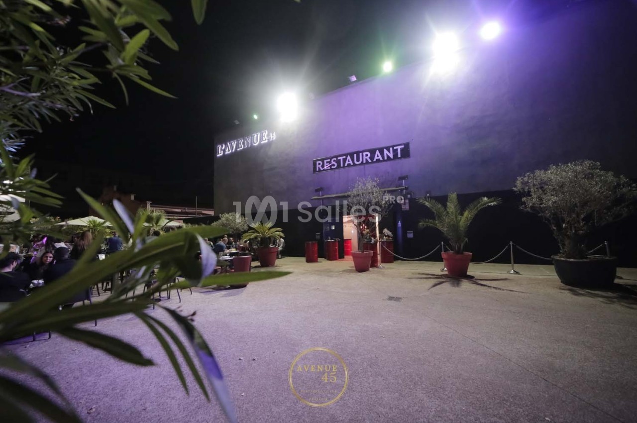 Façade d'un restaurant éclairé de nuit, entourée de plantes en pots sur une terrasse spacieuse.